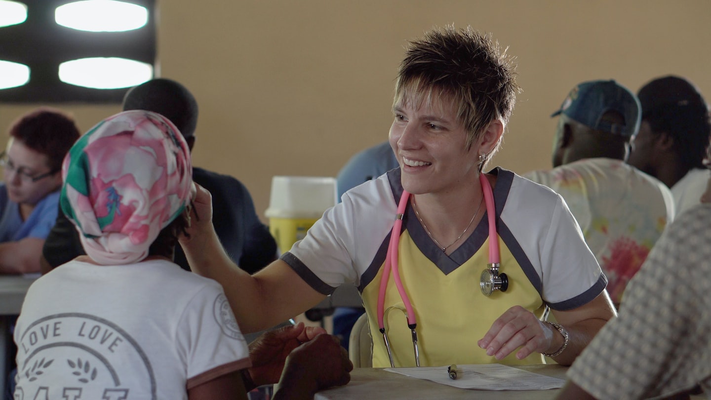 A nurse in yellow scrubs smiles at a patient seated at a table.